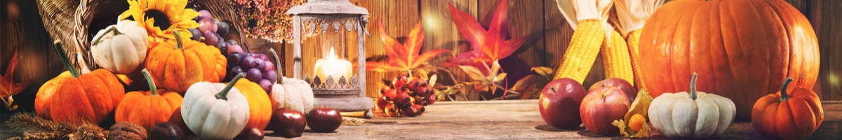 Orange und weiße Kürbisse, Äpfel, Nüsse, Maiskolben, Weintrauben und herbstliche rote Ahornblätter, gemütlich dekoriert auf einem Holztisch. Eine Kerze spendet warmes Licht.